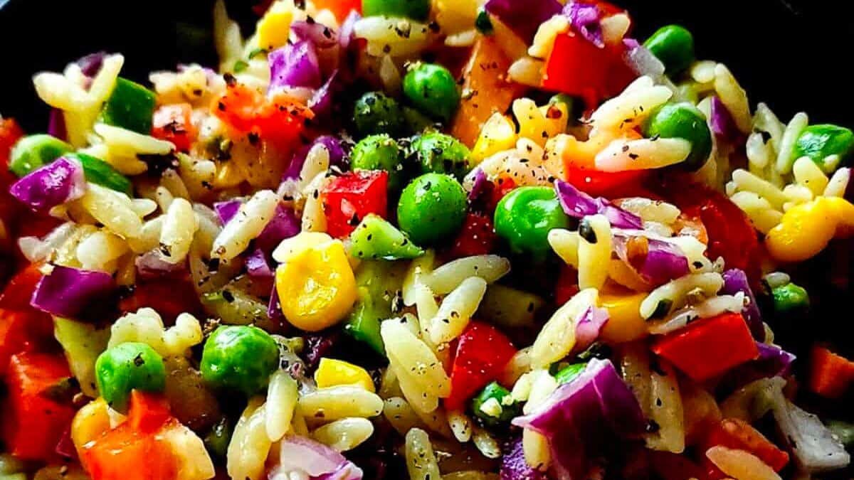 A vibrant salad featuring orzo pasta, red onions, bell peppers, green peas, corn, and purple cabbage, sprinkled with black pepper. The colorful ingredients create a visually appealing and appetizing dish.
