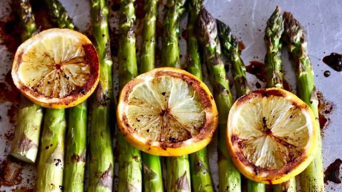Grilled asparagus spears topped with slices of roasted lemon, placed on a metal baking sheet. The lemons are golden brown, adding a vibrant touch to the green asparagus.