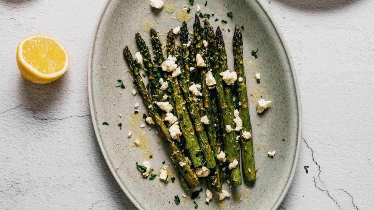 A ceramic oval plate with roasted asparagus topped with crumbled feta cheese and herbs. A lemon half is beside the plate on a light gray surface.