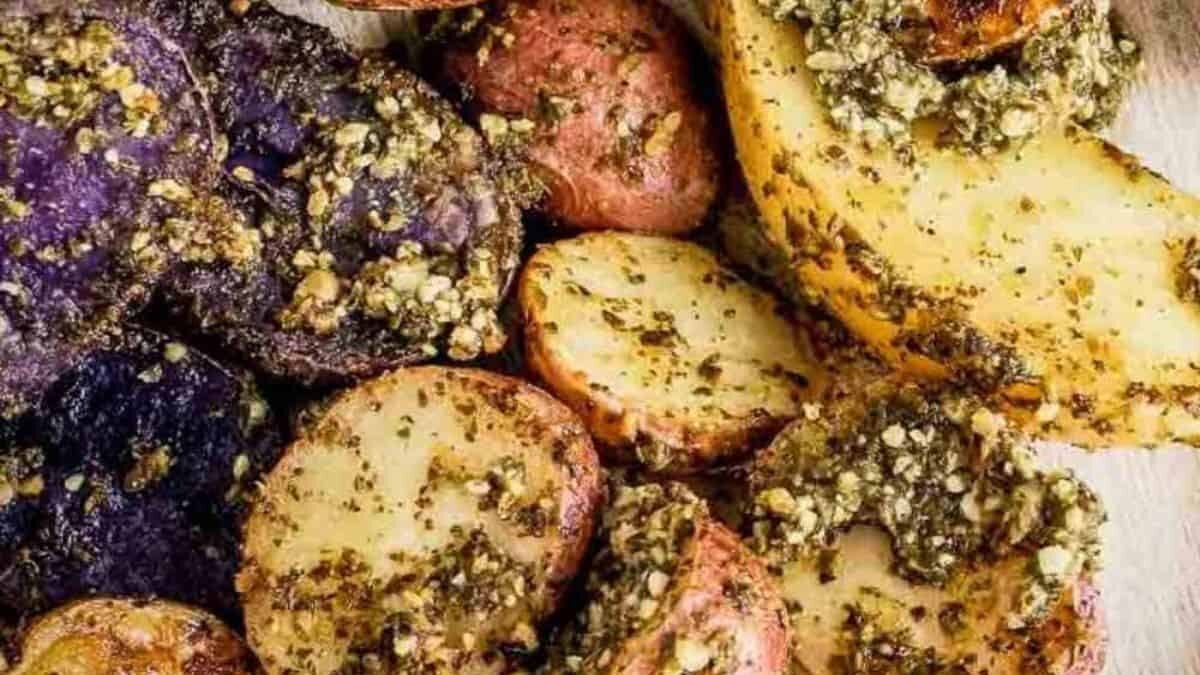 A close-up shot of assorted roasted potatoes, featuring red, yellow, and purple varieties. They are seasoned with a green pesto, providing a textured and flavorful coating. The potatoes are slightly golden, indicating a crispy, well-cooked finish.