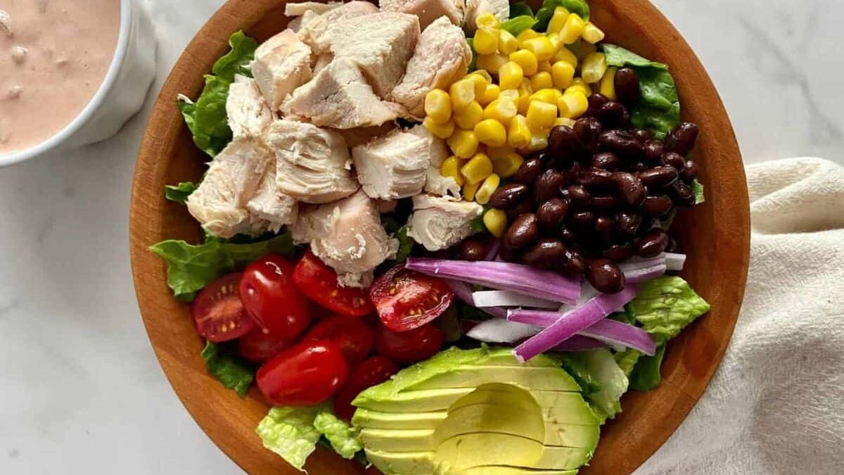 A wooden bowl filled with lettuce, diced chicken, corn, black beans, sliced red onion, cherry tomatoes, and avocado slices. A small bowl with sauce is on the side. The bowl is on a light marble surface.