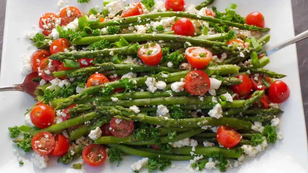 A rectangular plate filled with a fresh salad of asparagus, cherry tomatoes, and crumbled feta cheese, garnished with parsley. Two serving utensils are placed on opposite sides of the dish.