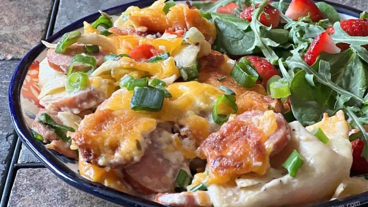 A plate of cheesy pierogi casserole topped with green onions, accompanied by a fresh salad with spinach, strawberries, and cherry tomatoes.