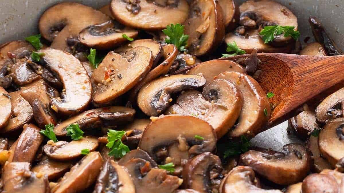 Sautéed mushrooms in a pan, garnished with fresh parsley. A wooden spoon is stirring the sliced mushrooms, which appear golden brown with bits of minced garlic and seasonings visible.