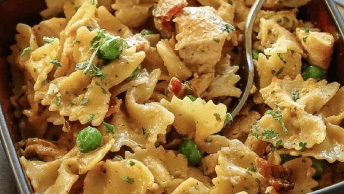 A close-up of bow-tie pasta mixed with pieces of chicken, green peas, bacon, and parsley in a creamy sauce. A spoon is placed in the dish.