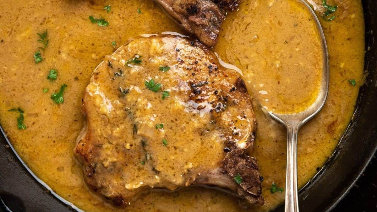 Pork chops in a creamy mustard sauce with herbs are served in a dark skillet. A spoon rests in the sauce, and fresh parsley is sprinkled on top for garnish.