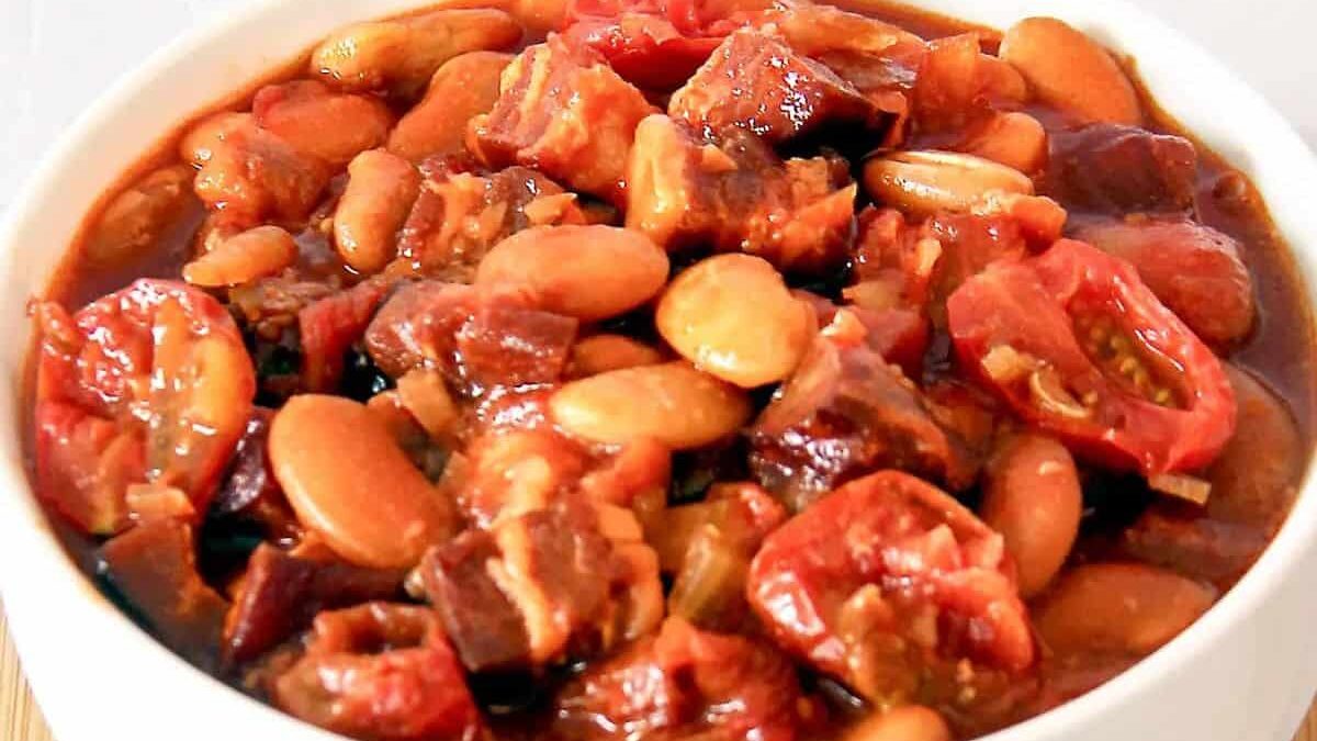 A bowl of baked beans with pieces of meat and tomatoes in a rich, thick sauce. The dish is garnished with sliced tomatoes and appears hearty and savory.