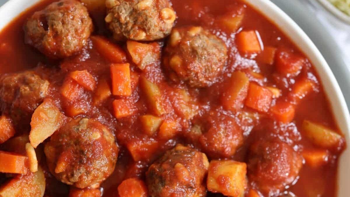 A bowl filled with meatballs in a rich tomato sauce, garnished with diced carrots and potatoes.