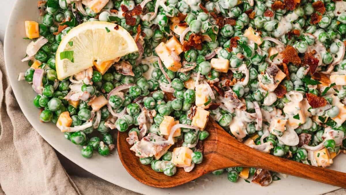 A close-up of a creamy pea salad with diced cheese, crispy bacon bits, chopped red onion, and fresh herbs, garnished with a lemon slice. The salad is served on a white plate with a wooden spoon, set on a beige cloth.