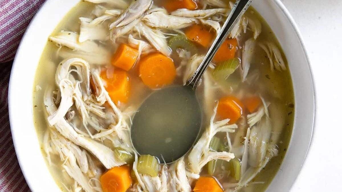 A bowl of chicken soup with shredded chicken, sliced carrots, and celery in clear broth, with a metal spoon resting inside the bowl.