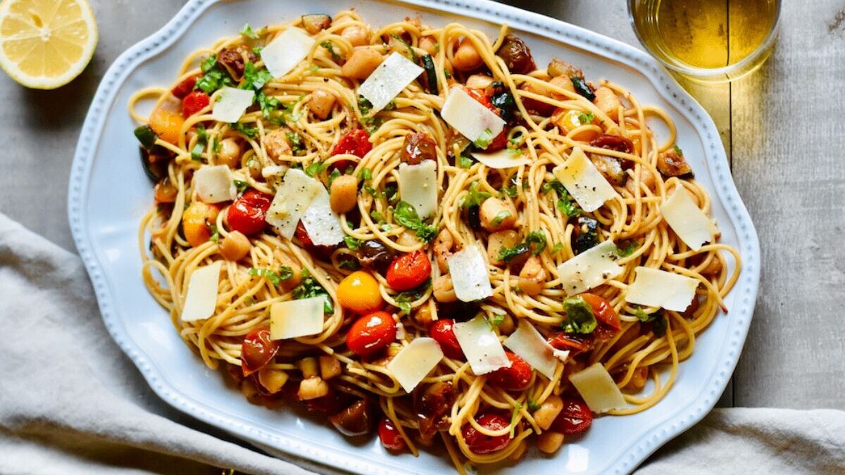 A plate of spaghetti with cherry tomatoes, sliced mushrooms, chickpeas, and spinach, topped with cheese shavings. A lemon wedge and a glass of white wine sit nearby on a wooden table.