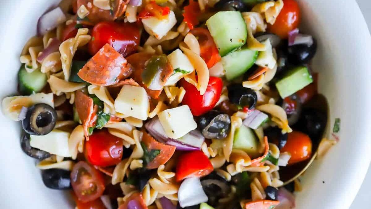 A bowl of pasta salad with spiral noodles, cherry tomatoes, cucumber chunks, black olives, mozzarella cubes, red onion, and slices of pepperoni, garnished with herbs.
