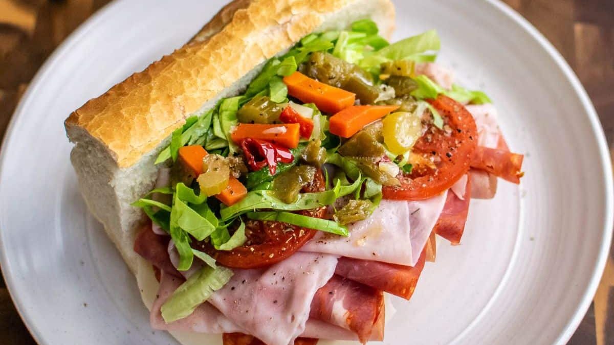 A sub sandwich on a white plate, filled with layers of ham, turkey, salami, lettuce, tomatoes, and mixed pickled vegetables on a crusty bread roll, garnished with black pepper.