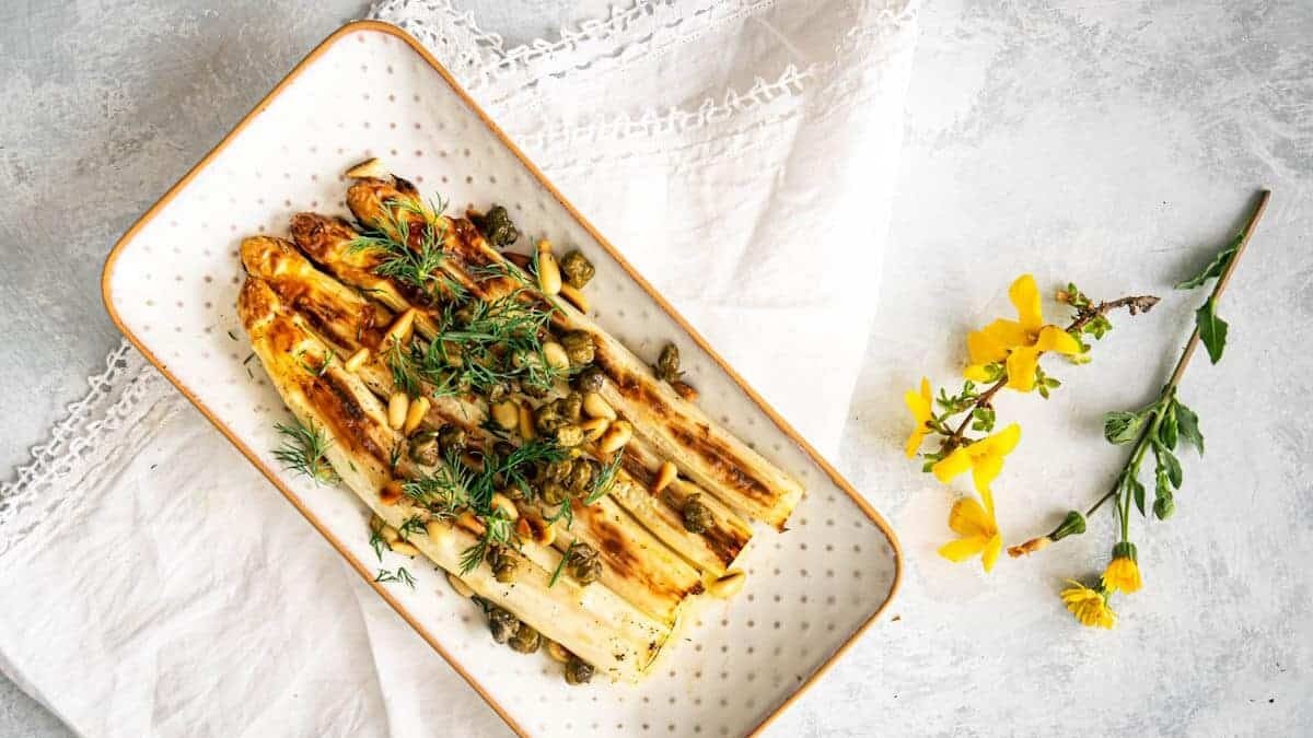 A rectangular white dish with a gold rim holds a serving of grilled white asparagus garnished with fresh dill, capers, and pine nuts. The dish is placed on a white cloth with lace edging, and a small sprig of yellow flowers lies beside it on a light gray, textured background.