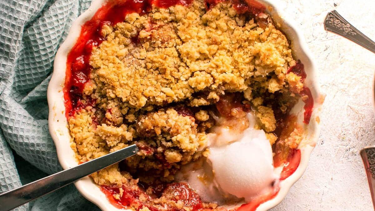 A close-up of a fruit crumble dessert in a white dish. The crumble has a golden-brown topping with visible chunks of fruit and a scoop of melting vanilla ice cream on one side. A spoon is placed inside the dish, and a light green textured cloth is nearby.