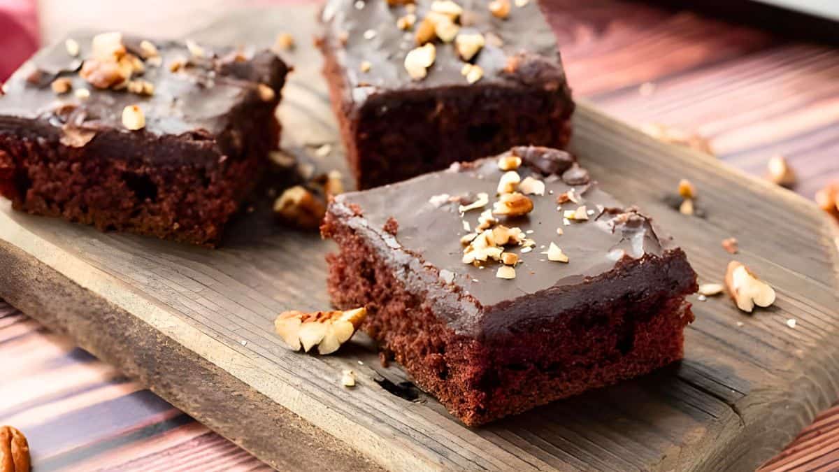 Three chocolate brownies topped with chopped nuts sit on a wooden board. The brownies have a glossy chocolate glaze, and scattered nuts surround the board on a wooden table.
