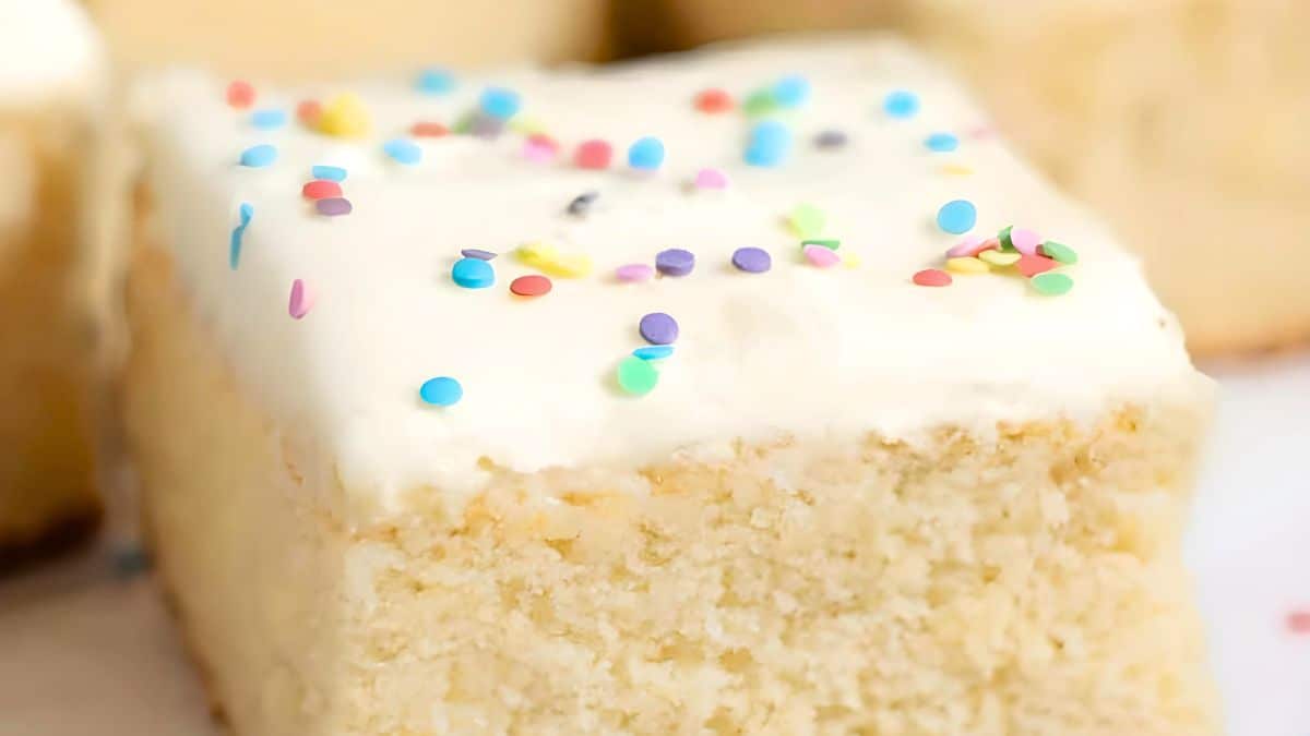 A slice of fluffy vanilla cake topped with creamy white frosting and sprinkled with colorful confetti sprinkles. The cake has a light, airy texture and looks moist and delicious.