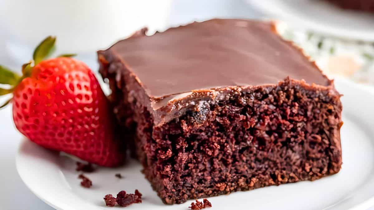 A slice of rich chocolate cake with a smooth, glossy chocolate icing sits on a white plate. Next to the cake is a fresh strawberry, adding a pop of red color to the scene.