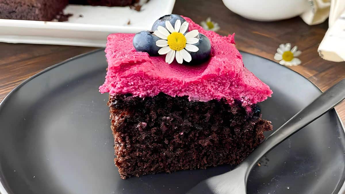 A slice of chocolate cake topped with vibrant pink frosting, adorned with fresh blueberries and a small daisy flower, served on a black plate with a fork on the side. A partial view of the larger cake is in the background.