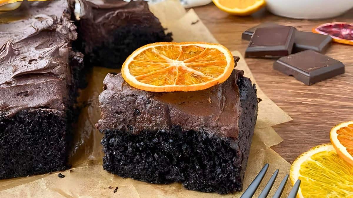 A piece of moist chocolate cake with rich frosting, topped with a dried orange slice, is placed on parchment paper. Slices of fresh and dried oranges and pieces of dark chocolate surround the cake. A fork rests nearby.