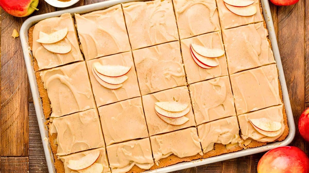 A rectangular sheet pan holds frosted apple bars cut into squares. The frosting is light brown and smooth. Several squares are topped with thin apple slices arranged in a fan shape. Surrounding the pan are whole apples, some partially visible.