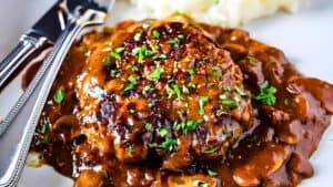 A juicy Salisbury steak covered in rich brown gravy with mushrooms, garnished with chopped parsley. A knife and fork are placed beside it, and a portion of mashed potatoes is visible in the background.