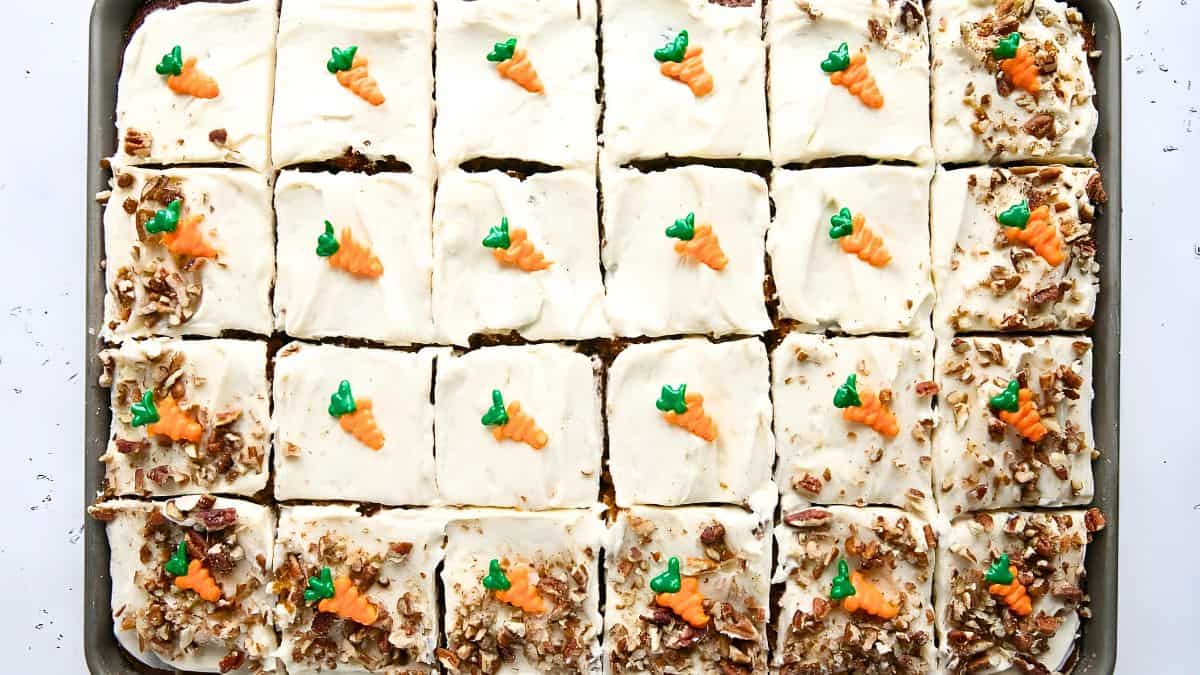 A tray of frosted carrot cake squares topped with small icing carrots. Some squares are sprinkled with chopped pecans. The cake is arranged in a grid pattern.