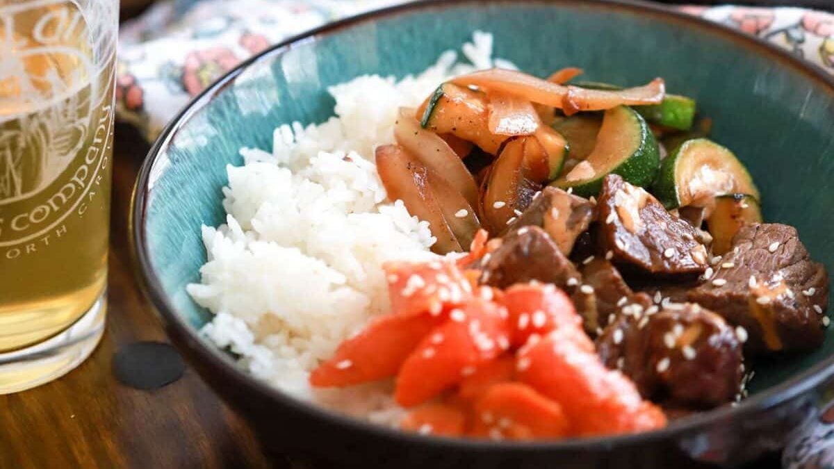A bowl filled with white rice, sautéed vegetables, and chunks of beef topped with sesame seeds, with a glass of light beer nearby on a wooden table.