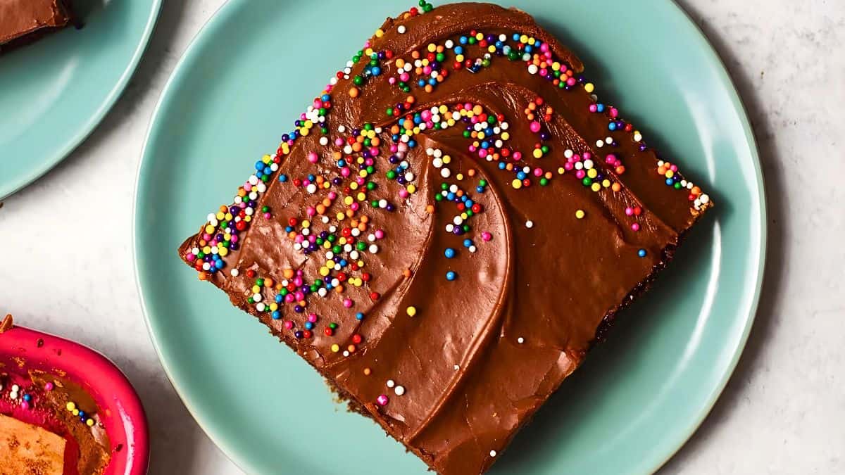A slice of chocolate cake with chocolate frosting and colorful sprinkles on a teal plate. The cake is square, with smooth frosting and scattered sprinkles adding a festive touch. Another partially visible plate with dessert appears in the corner.