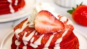 A stack of red velvet pancakes drizzled with white icing, topped with a dollop of whipped cream and a sliced strawberry. A whole strawberry is on the side.