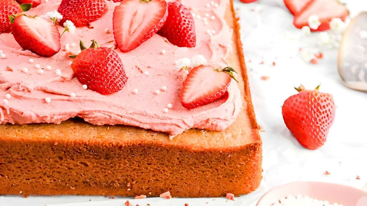 A close-up of a rectangular cake topped with pink frosting and whole and sliced strawberries. The cake sits on a white surface with a few strawberries scattered around. Small white sprinkles decorate the frosting.