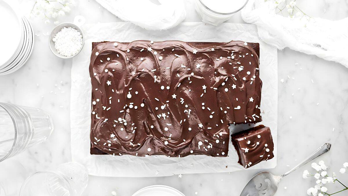A rectangular chocolate sheet cake with swirled frosting and white sprinkles sits on a marble surface. One piece is cut and placed next to the cake. Nearby are white dishes, a glass, and small flowers scattered around.