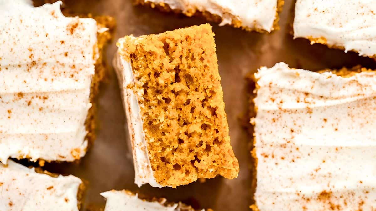 Close-up of frosted pumpkin bars on a wooden surface. The bars are topped with creamy white frosting and lightly sprinkled with cinnamon. One bar is positioned vertically, showcasing its moist texture.
