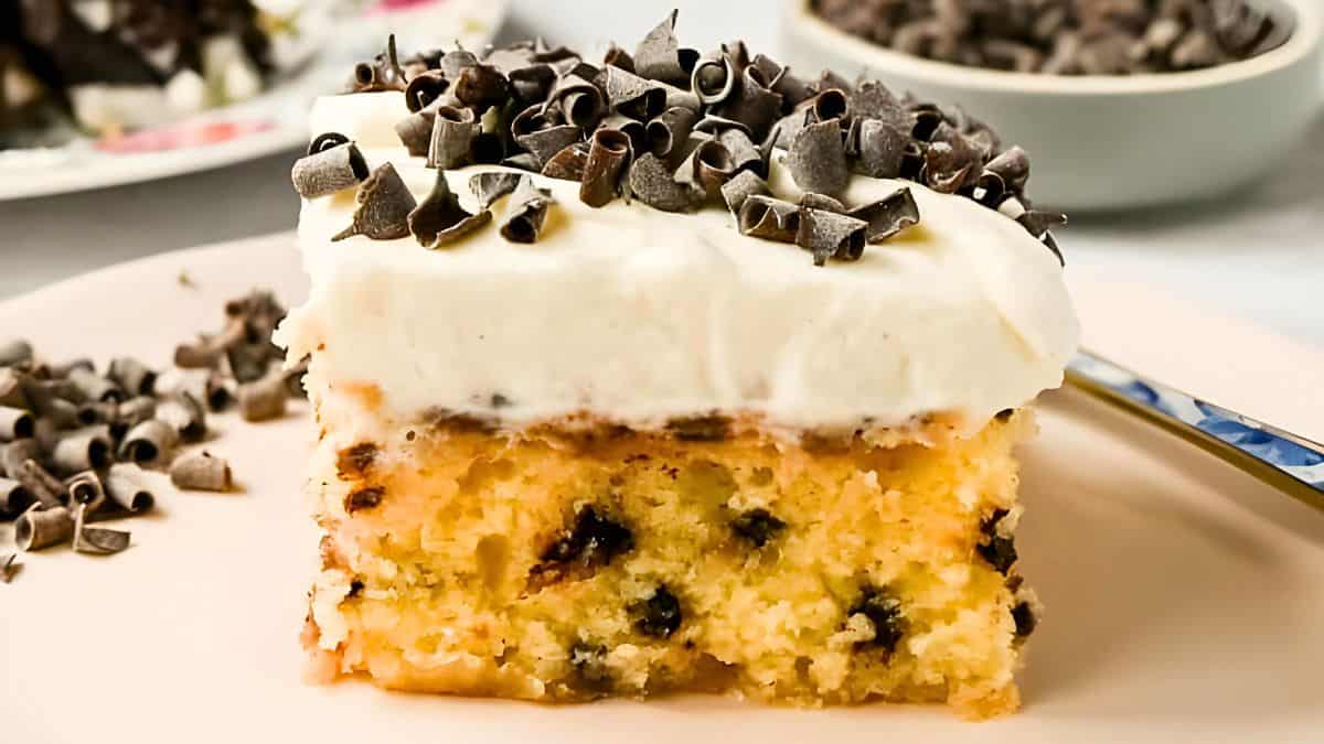 A slice of moist vanilla cake topped with creamy white frosting and garnished with dark chocolate shavings sits on a light pink plate. A fork rests nearby, and there is a bowl of chocolate shavings in the background.