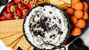 A platter featuring a central bowl of creamy dip sprinkled with crushed cookies. Surrounding it are strawberries, pretzels, apple slices, graham crackers, and round cookies, offering a variety of snacks for dipping.