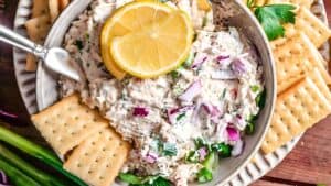 A bowl of creamy tuna salad garnished with lemon slices and chopped red onions, surrounded by square crackers on a plate. Fresh herbs and green onions accompany the dish. A small serving spoon is placed in the bowl.