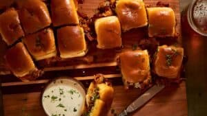 A wooden board with pulled pork sliders topped with shiny buns, garnished with herbs. A small bowl of creamy dipping sauce is nearby, alongside a knife with sandwich filling. A glass of beer is partially visible on the right.