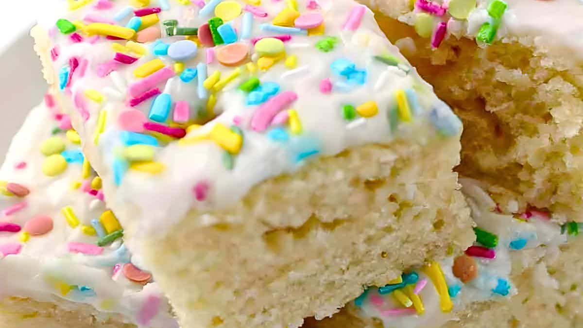 Close-up of frosted cake squares covered with colorful sprinkles, including circles and rods in pink, yellow, orange, green, and blue. The texture of the cake appears soft and fluffy.