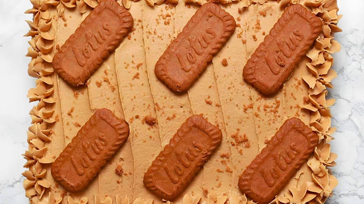 A cake topped with a smooth, light brown frosting, adorned with six rectangular Lotus biscuits and crumbled biscuit pieces along the edges. The sides are decorated with piped swirls of frosting.