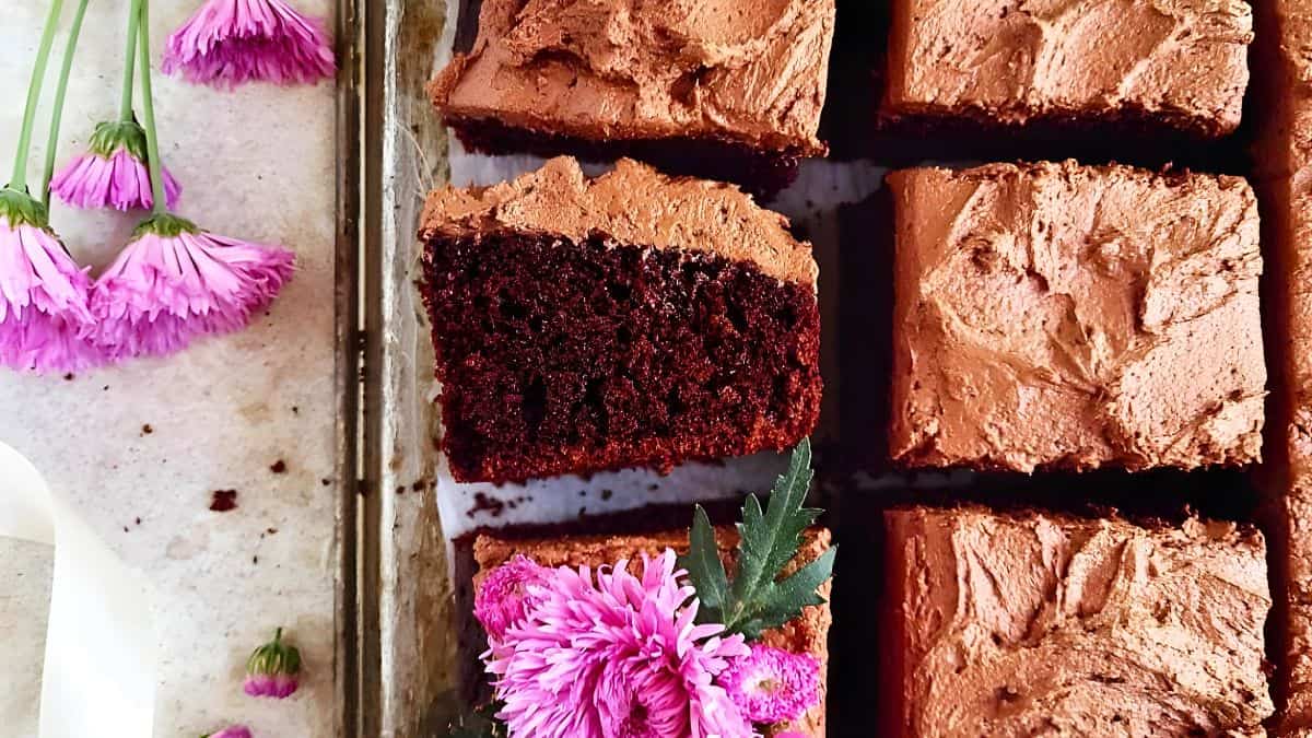 Slices of chocolate cake with creamy frosting are arranged neatly. One slice is turned slightly to reveal its rich texture. Fresh pink flowers are placed decoratively beside the cake on a marble surface.
