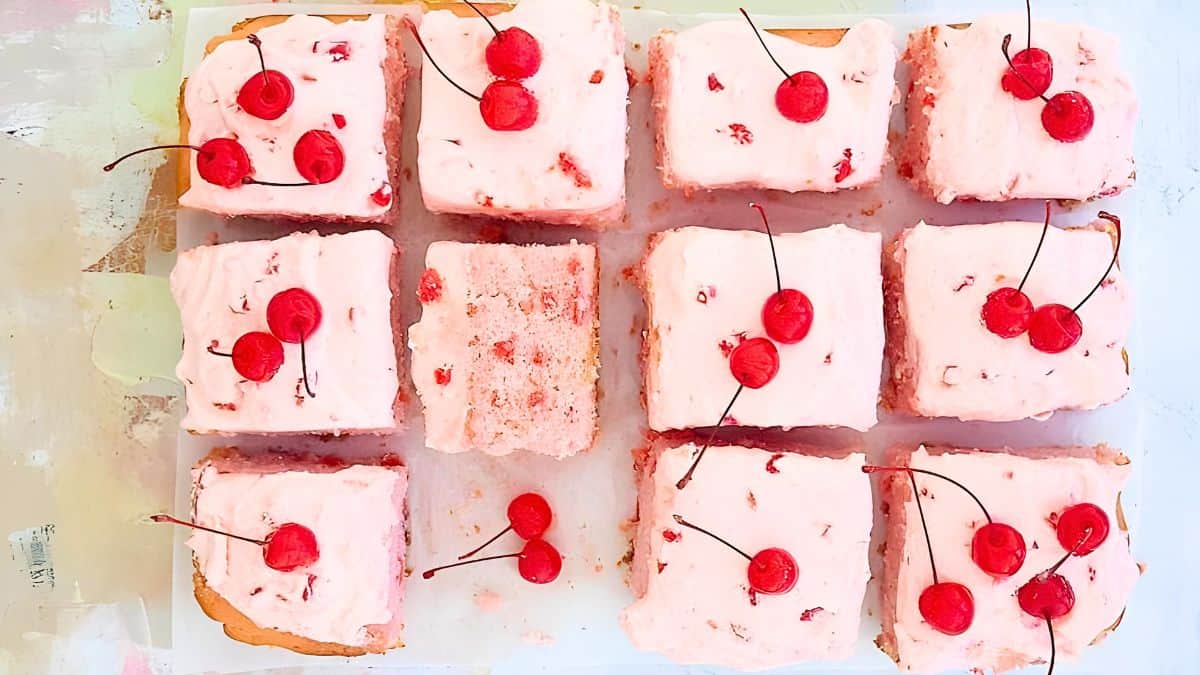 Twelve square pink frosted cakes topped with red cherries. One piece is slightly taken, showing a fluffy interior. The cakes are arranged in a grid on a light surface.