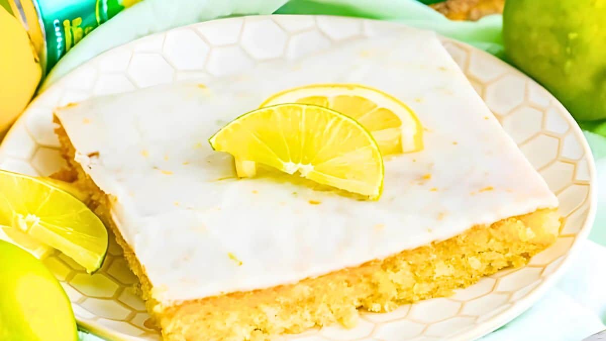 A square piece of lemon cake topped with white icing and lemon slices sits on a patterned plate. Green apples and lemons surround the plate, enhancing the fresh and citrusy presentation.