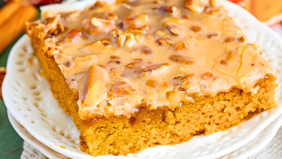 A close-up of a slice of pumpkin cake on a white plate. The cake is topped with a creamy, nut-filled glaze, showcasing a rich amber color and moist texture. The decorative plate edge is visible beneath the dessert.
