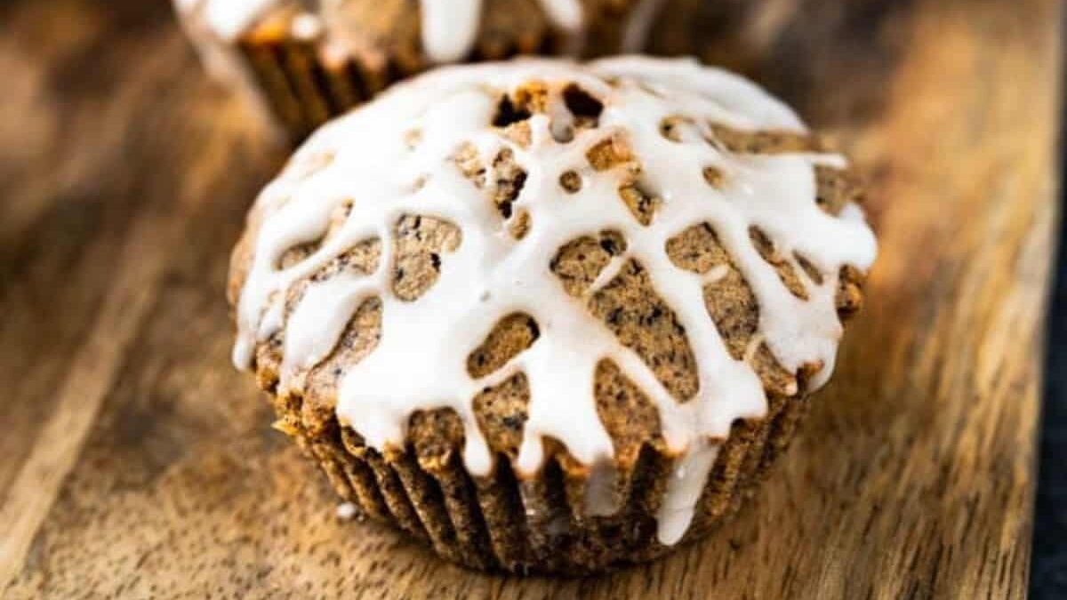 A close-up of a muffin on a wooden surface, topped with a white glaze drizzled in a zigzag pattern. The muffin has a slightly textured, golden-brown appearance, suggesting it's freshly baked.