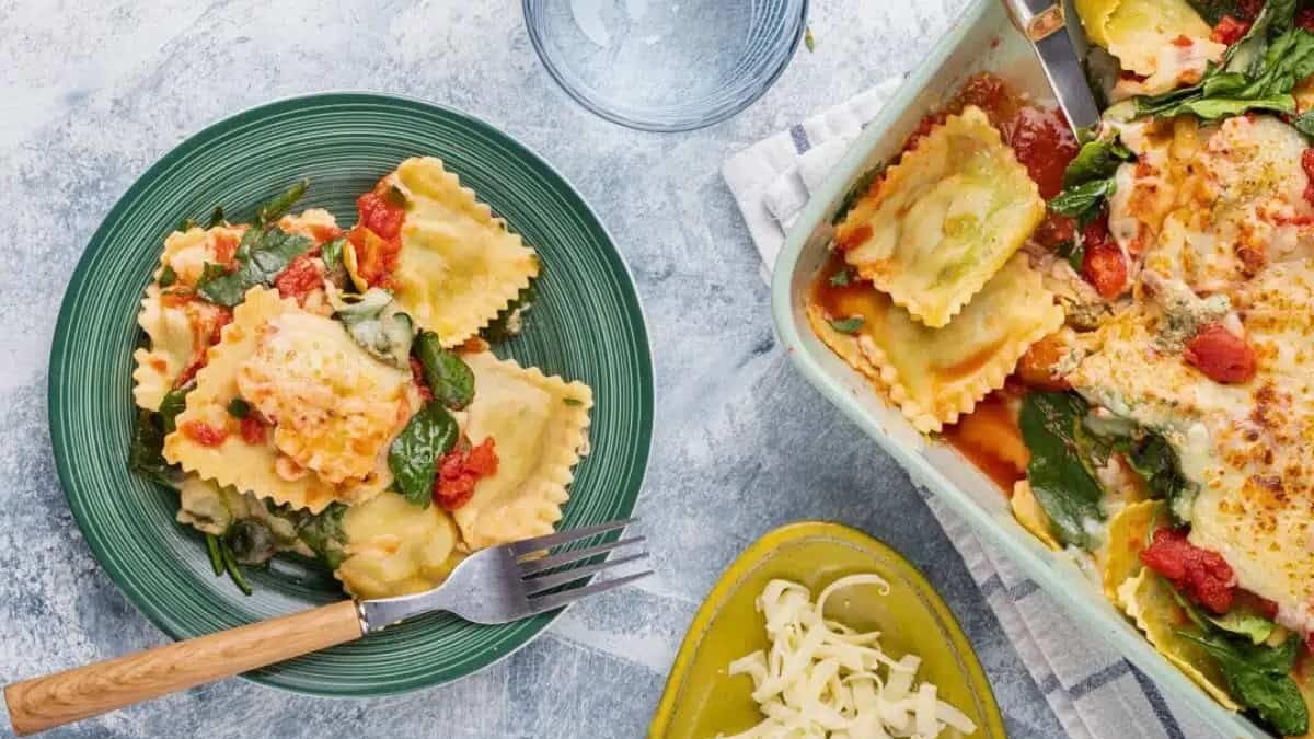 A green plate with cheese ravioli topped with spinach and tomato sauce sits next to a fork. Nearby, a casserole dish filled with ravioli, vegetables, and cheese, a bowl of grated cheese, and an empty glass are placed on a textured surface.