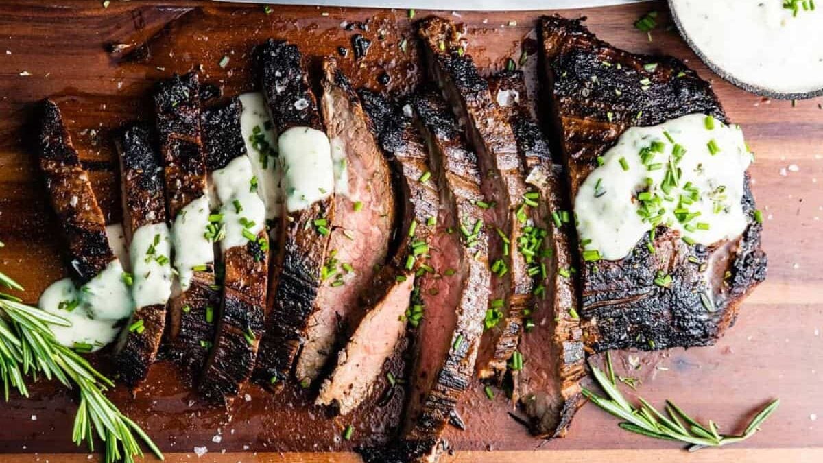 Sliced grilled steak topped with creamy white sauce and chopped chives on a wooden cutting board, garnished with rosemary sprigs. A large knife and a small bowl of sauce are also visible.