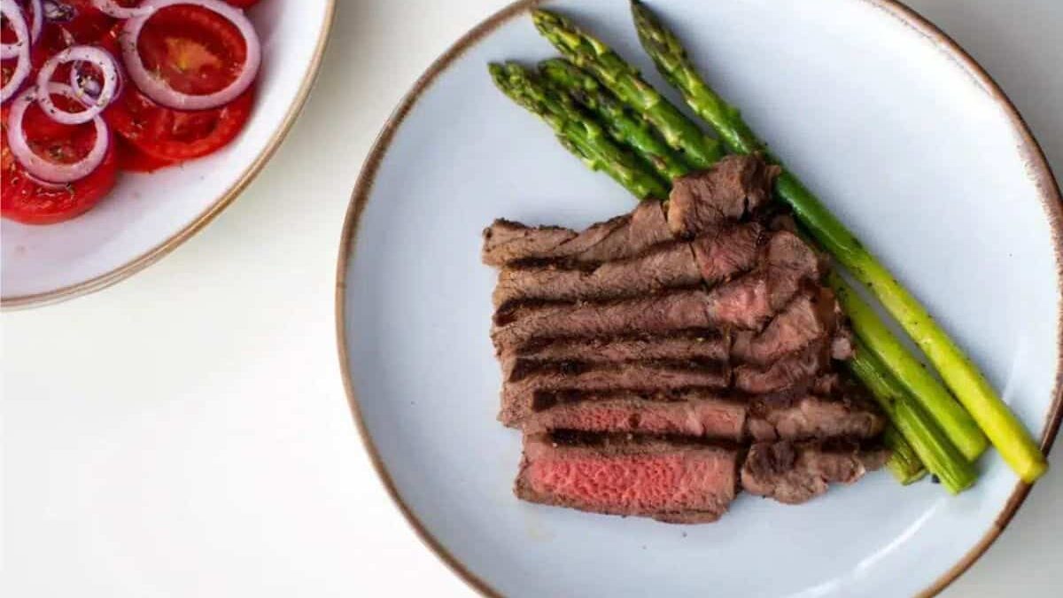A plate with slices of medium-rare steak and asparagus spears. Next to it is another plate with sliced tomatoes topped with red onion rings.