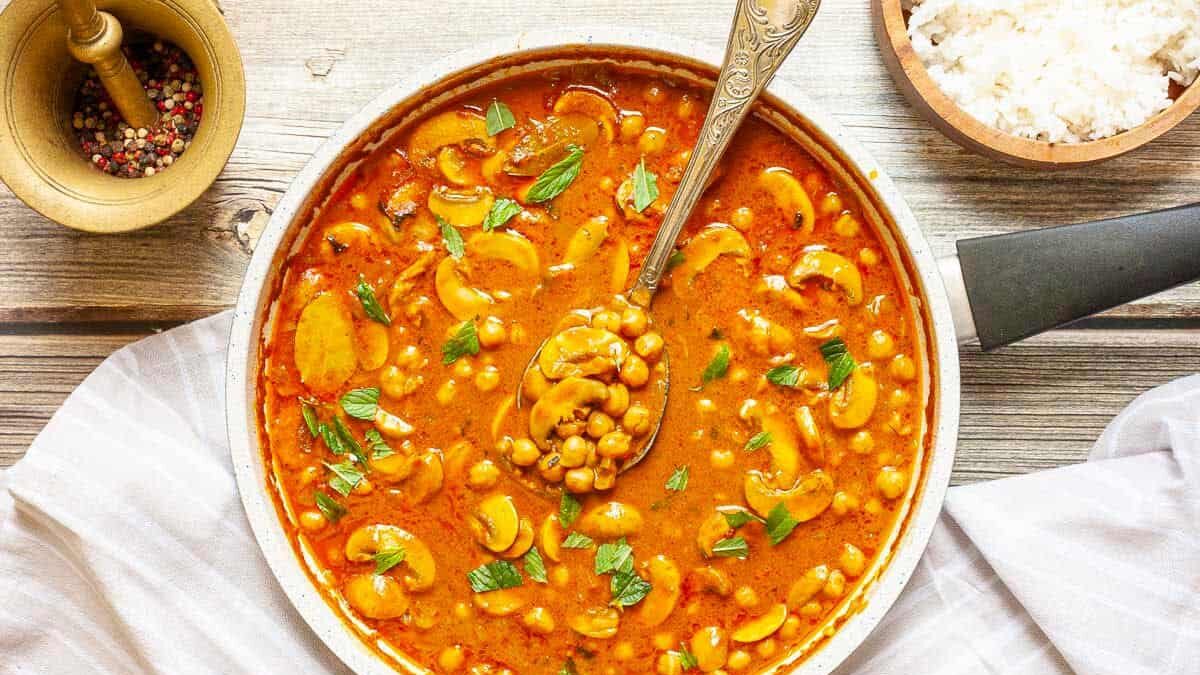 A skillet filled with creamy curry featuring chickpeas, cashews, and garnished with fresh herbs. A spoon rests in the curry. A small bowl of rice and a pepper grinder are nearby on a wooden surface with a white cloth.