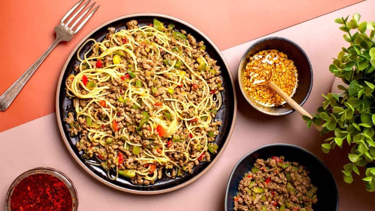 A plate of spaghetti with minced meat, red and green peppers, and zucchini, served with a side of extra toppings and a bowl of red sauce. A fork and a small plant decorate the setting on a two-tone table.