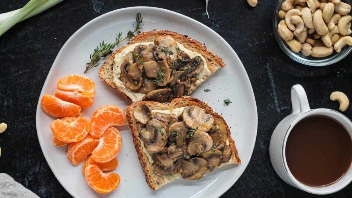 A plate with two slices of toast topped with mushrooms, a peeled and segmented mandarin orange, and a cup of black coffee. A bowl of cashews and scattered cashews are nearby on a dark surface.